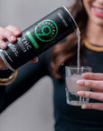 A person holds a can of Crescent 9 Raspberry Lime THC Seltzer and pours it into a small clear shot glass. The background is a softly blurred indoor setting.