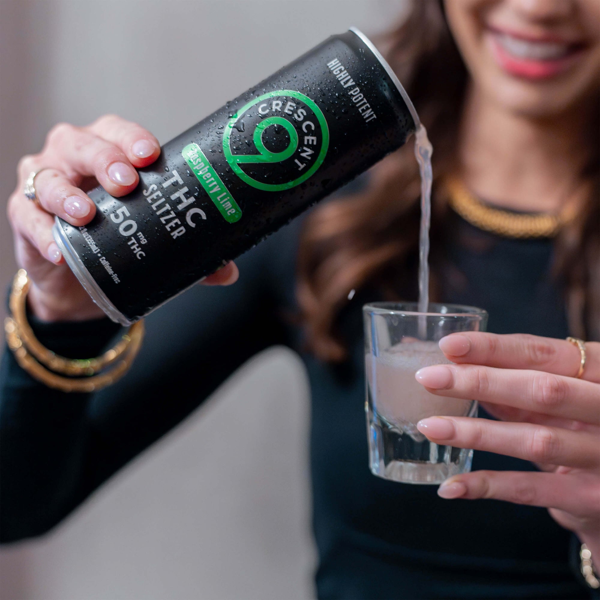 A person holds a can of Crescent 9 Raspberry Lime THC Seltzer and pours it into a small clear shot glass. The background is a softly blurred indoor setting.