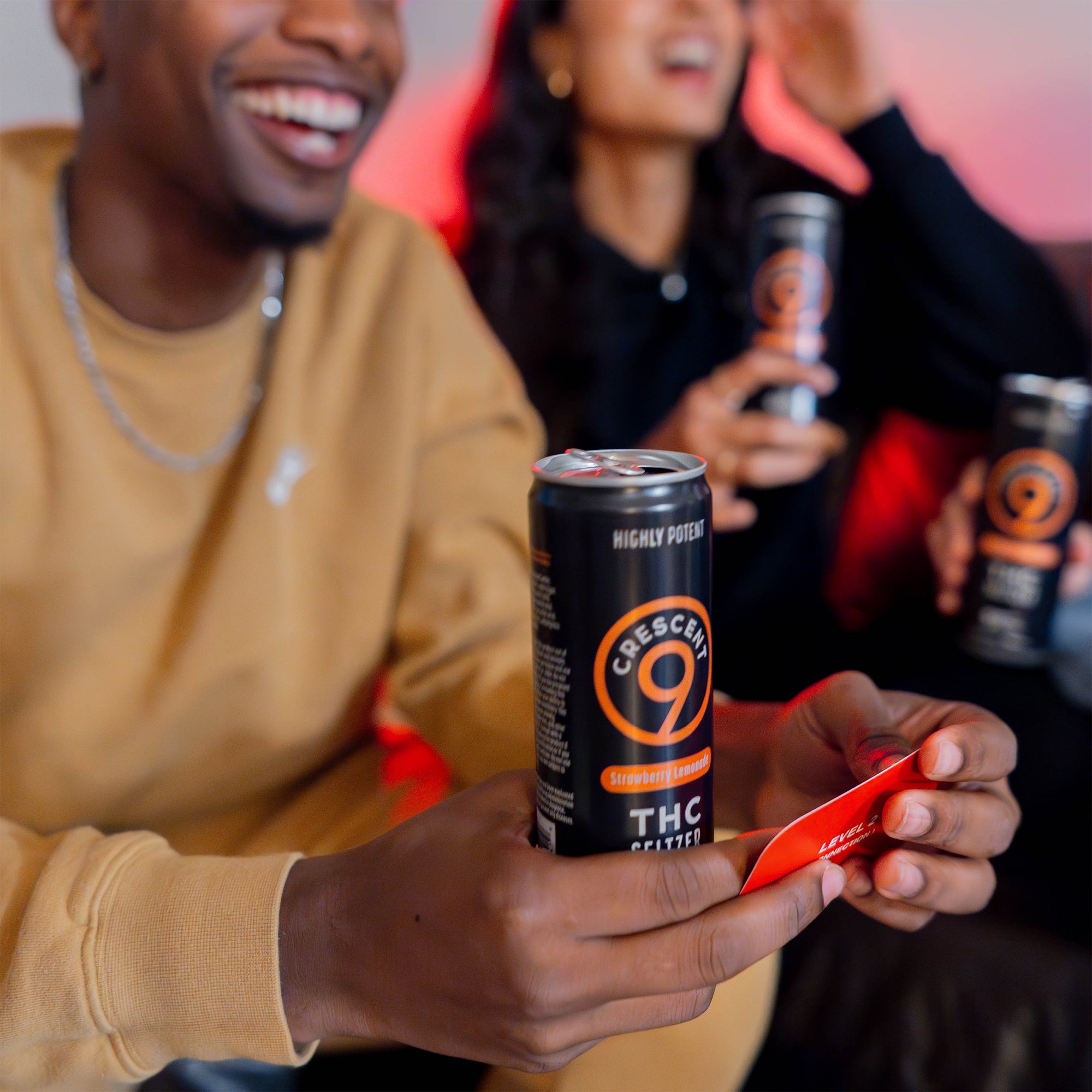 A man in a tan sweatshirt holds a can of Crescent 9 THC seltzer, with a red card in his hand, while a woman in a black top smiles in the background holding another can.